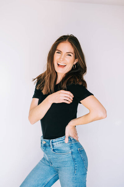 Indoor portrait of beautiful laughing woman, wearing black t-shirt and high waist jeans, posing on white background