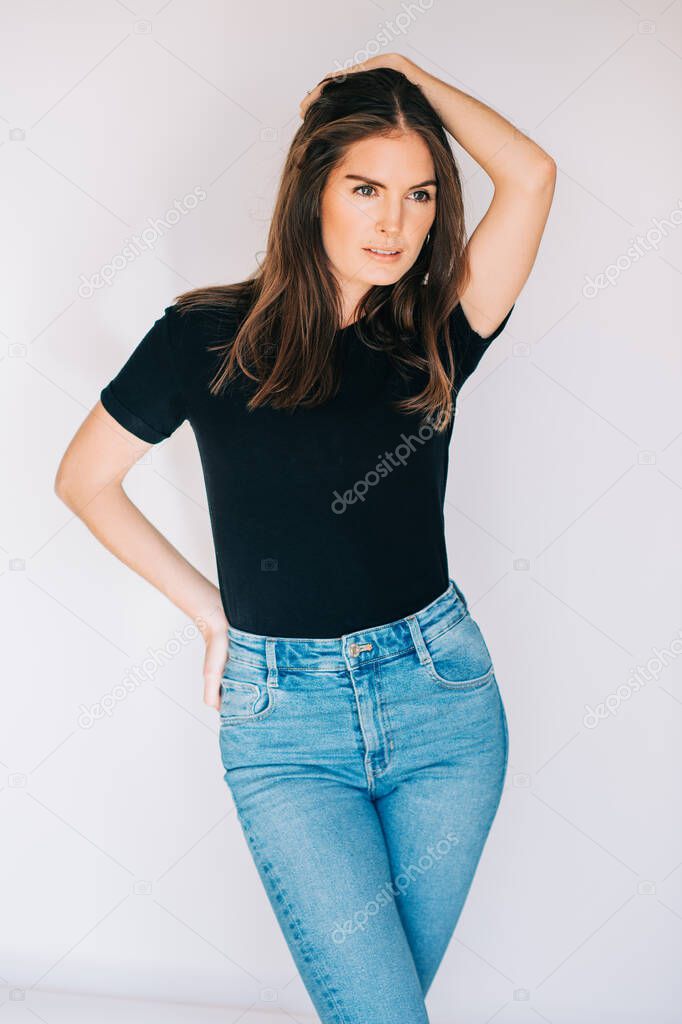 Indoor portrait of beautiful young woman, wearing black t-shirt and high waist jeans, posing on white background, holding hand on head
