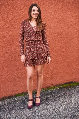 Açık havada uzun düz kahverengi saçlı güzel bir kadının portresi. Terracotta duvarının arka planında poz veriyor. Mini elbise giyiyor.