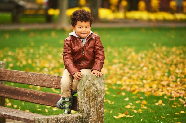 可爱的非洲男孩 穿着棕色皮夹克 公园里玩得开心 坐在长椅顶上的户外秋天肖像 — 图库照片