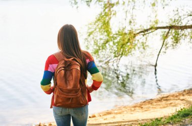 Güzel bir kadının açık hava portresi güzel bir günde nehirde dinleniyor, renkli bir kazak ve sırt çantası giyiyor.