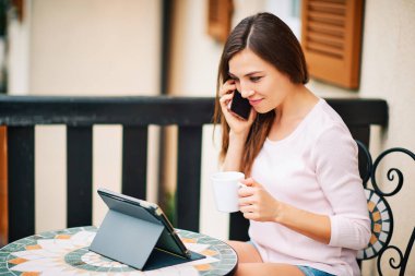 Balkonda çalışan, tablet kullanan, telefonla konuşan genç bir kadın.