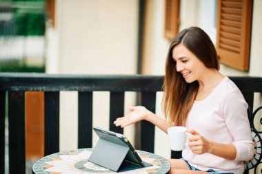 Balkonda çalışan, tablet tablet kullanan, elinde kahve tutan genç bir kadın.