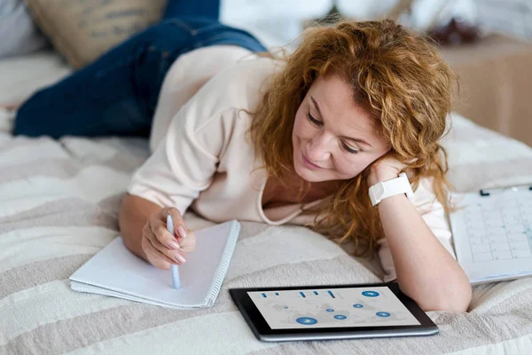 Donna sdraiata sul letto con tablet e appunti — Foto Stock