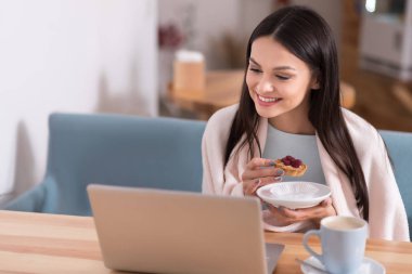 Laptop kullanan ve kafede pasta yiyen neşeli bir kadın..