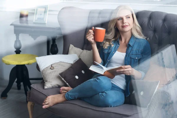Mladá blond žena sedící na gauči s knihou a pohár — Stock fotografie