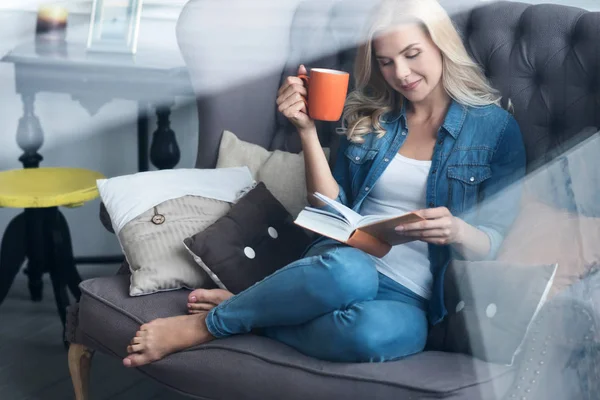 Mladá blond žena sedící na gauči s knihou a pohár — Stock fotografie