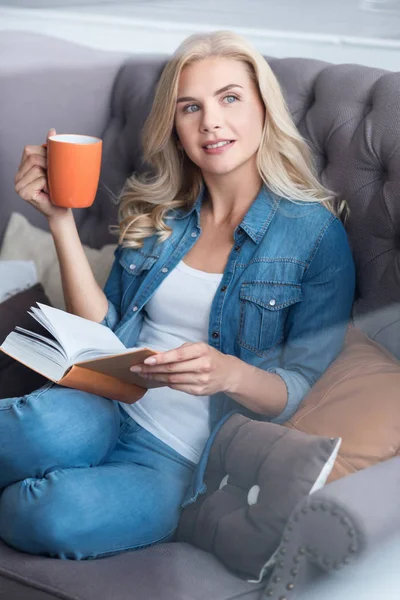 Fiatal szőke nő ül a kanapén könyv és kupa — Stock Fotó
