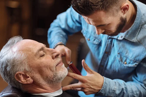 Красивый молодой парикмахер стрижет усы старшего клиента ножницами — стоковое фото
