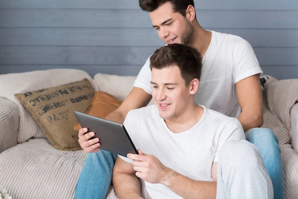 Relajada pareja gay descansando en el sofá y usando la tableta . —  Fotos de Stock