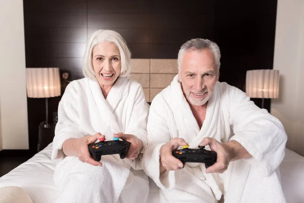 Alegre pareja de edad jugando con la consola de juegos en la cama — Foto de Stock