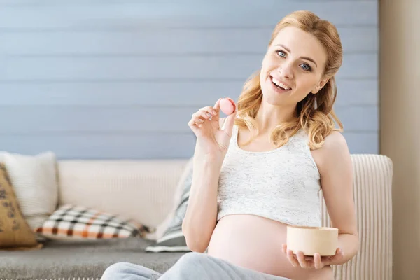 Pregnant woman holding macaroon on a sofa — Stock Photo, Image