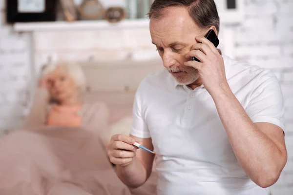 Hombre mayor llamando a emergencia mientras revisa el termómetro — Foto de Stock