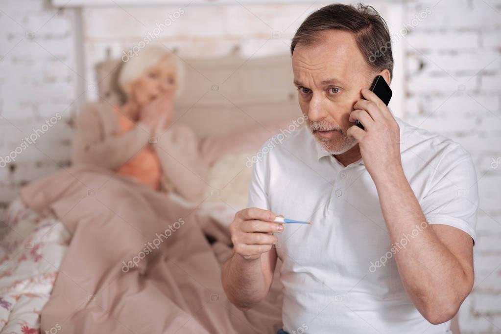 Come soon. Worried elderly man holding thermometer and calling emergency on background of his sick senior wife.