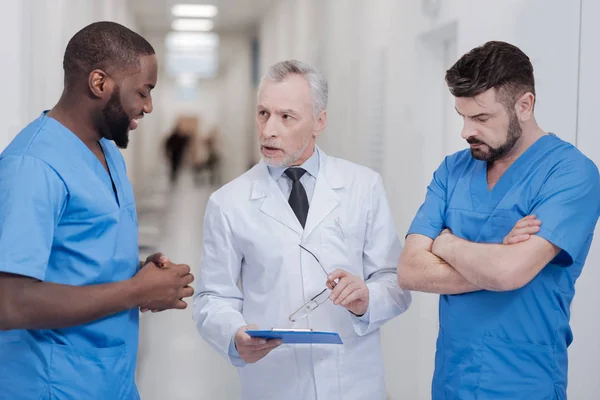 Praticante idoso rigoroso consultar jovens colegas no hospital — Fotografia de Stock