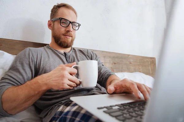 Cheerful man drinking coffee and working with laptop in bed — Stock Photo, Image