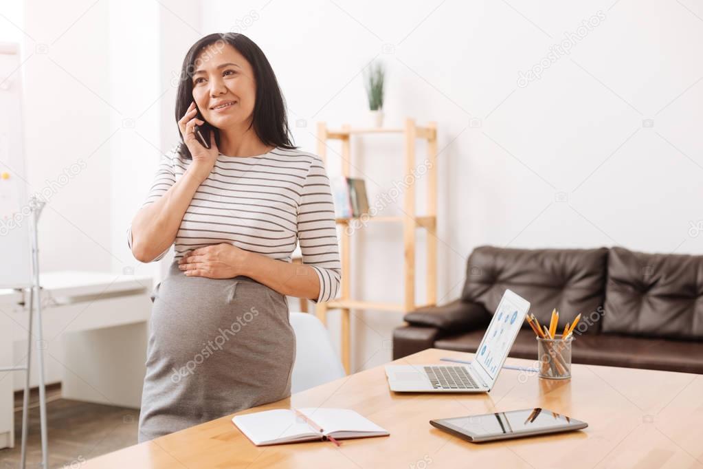 Call me soon. Delighted asian pregnant woman talking on phone and standing in the office while waiting for a maternity leave