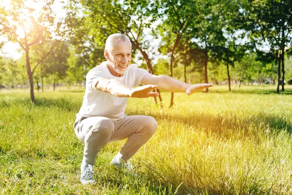 Позитивный пожилой мужчина делает приседания в парке — стоковое фото