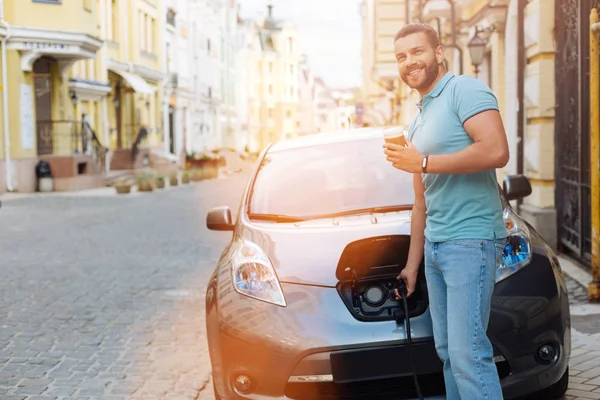 Bonito homem bebendo café enquanto carrega carro elétrico — Fotografia de Stock