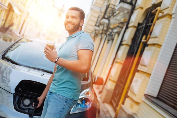 Молодой человек заряжает машину и пьет кофе. — стоковое фото
