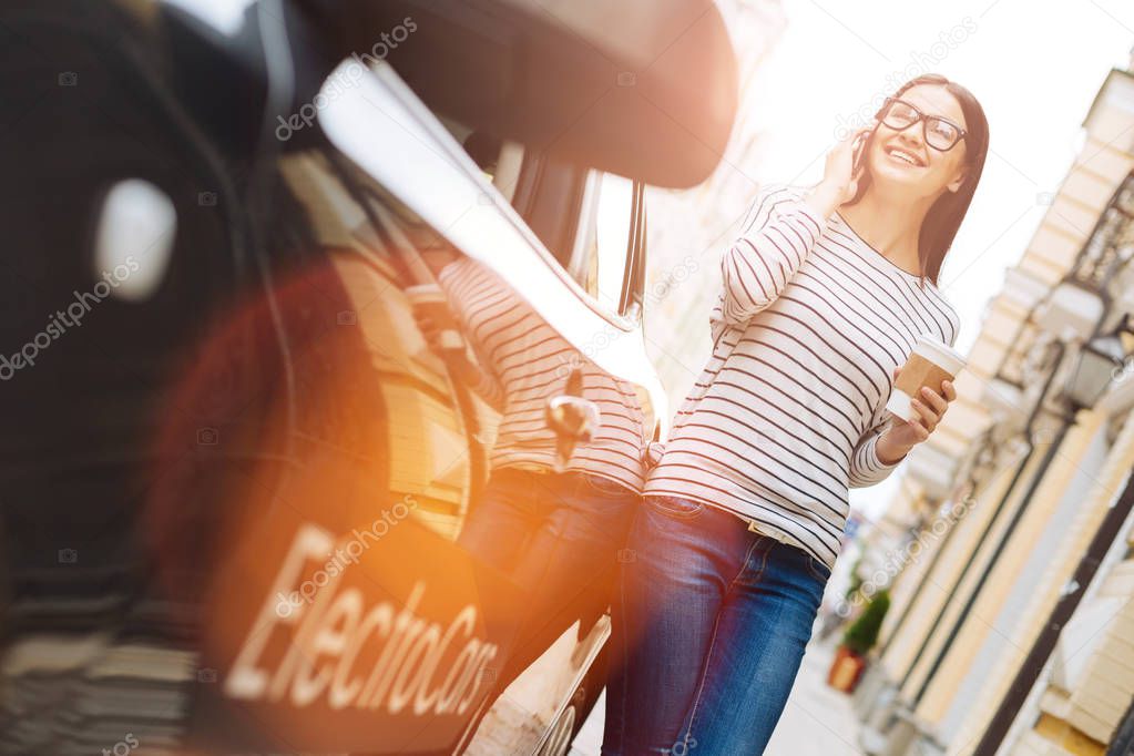 Pleasant conversation. Charming young woman in glasses holding a cup of coffee and talking on the phone while standing near her new electric car