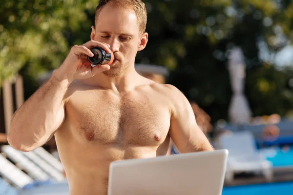 Sturdy man drinking fizzy drink while looking at laptop — Stock Photo, Image