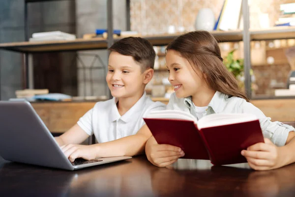 Little girl reading book while her brother playing on laptop