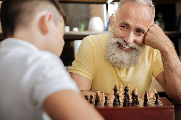 Hombre mayor alegre sonriendo a su nieto mientras juega al ajedrez — Foto de Stock