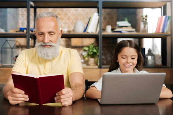 Agradable abuelo leyendo libro mientras que su nieta usando el ordenador portátil — Foto de Stock