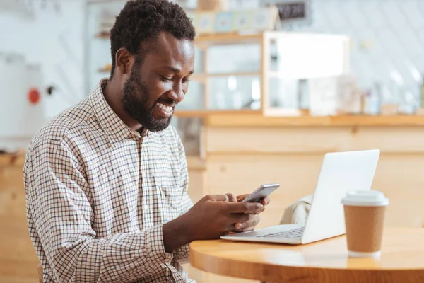 Feliz hombre leyendo el mensaje por teléfono mientras está sentado en la cafetería — Foto de Stock