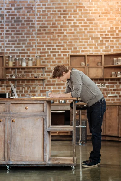 Όμορφος νεαρός άνδρας, καθιστώντας τις σημειώσεις στον πάγκο κουζίνας — Φωτογραφία Αρχείου