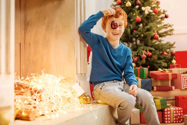 Chico jengibre emocionado jugando con la bola decorativa junto a la chimenea — Foto de Stock