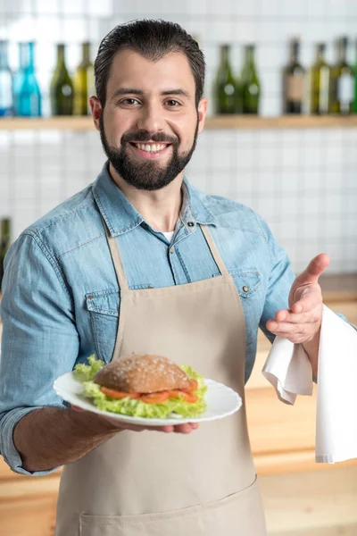 Freundlicher bärtiger Kellner, der lächelnd auf das leckere Sandwich zeigt — Stockfoto