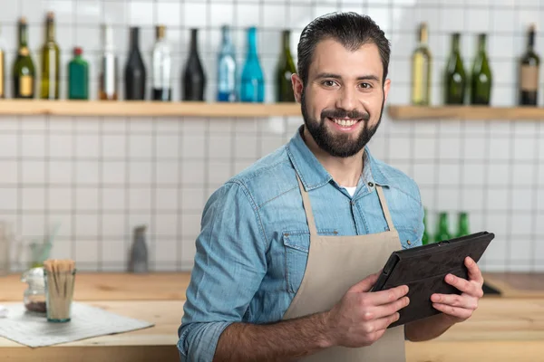 Alegre camarero barbudo sosteniendo una tableta moderna y sonriendo —  Fotos de Stock