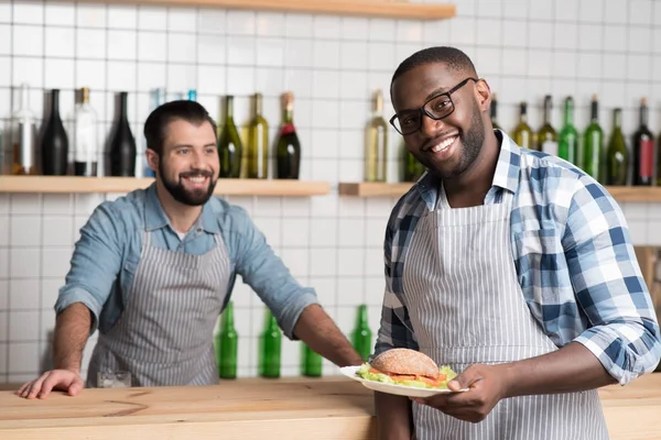 Joven camarero emocional sosteniendo un sándwich mientras su colega sonríe —  Fotos de Stock