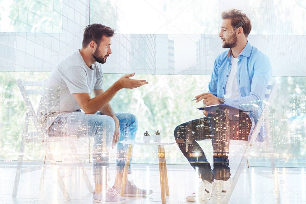 Being in trouble. Young handsome bearded man explaining his problems and raising right hand while psychologist making notes and listening to him carefully