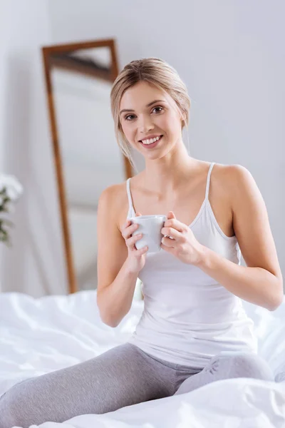 Mujer alegre arrodillada en la cama y sosteniendo una taza de café — Foto de Stock