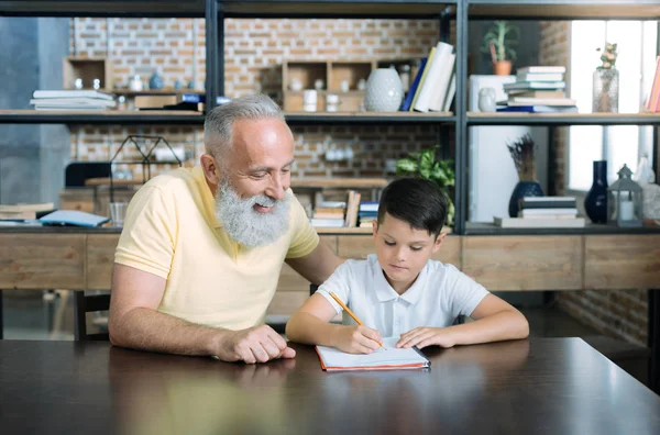 Extremadamente feliz anciano mirando a su nieto haciendo su tarea — Foto de Stock