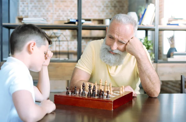 Amar al abuelo sonriendo mientras mira a su nieto pensando en el ajedrez — Foto de Stock