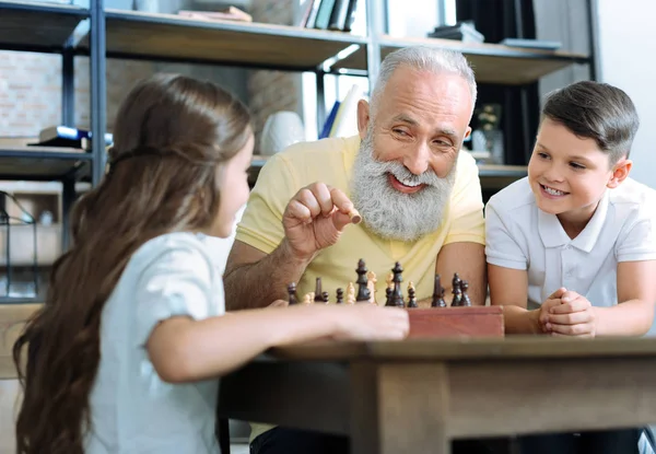 Любящий старший джентльмен улыбается, обучая внуков шахматам — стоковое фото