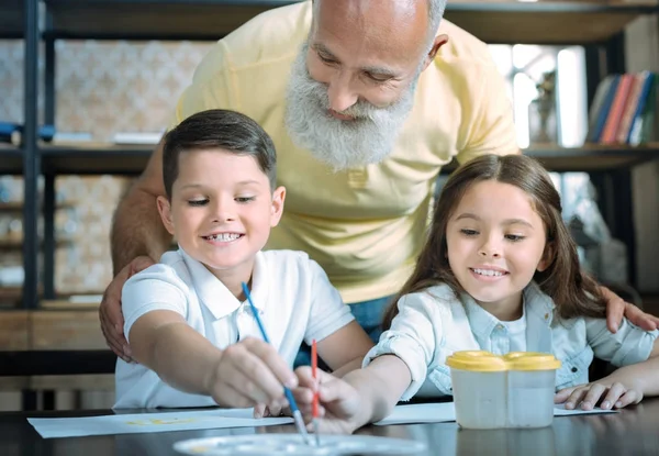 Vorsichtiger Opa schaut Kindern beim gemeinsamen Malen zu — Stockfoto