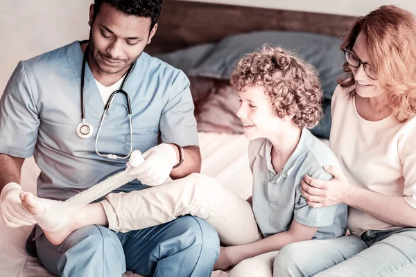 Niño radiante mirando al doctor vendándose el pie — Foto de Stock