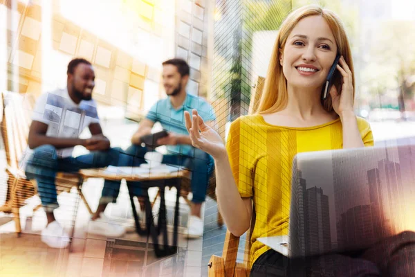 Joven mujer sonriente hablando por teléfono y sentada con un portátil — Foto de Stock