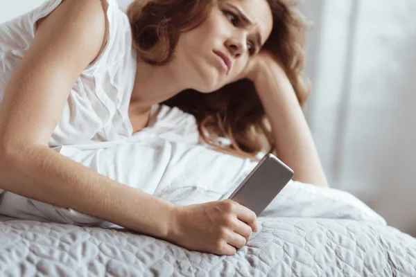 Mujer triste pensativa sosteniendo teléfono inteligente mientras está acostado en la cama —  Fotos de Stock