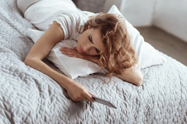 Brunette sedih berbaring di tempat tidur dengan smartphone di tangannya — Stok Foto