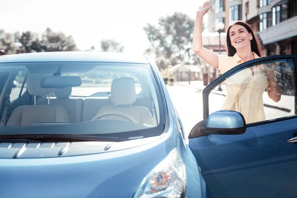 Zufriedene angenehme Frau, die neben dem Auto steht und die Hand hebt. — Stockfoto