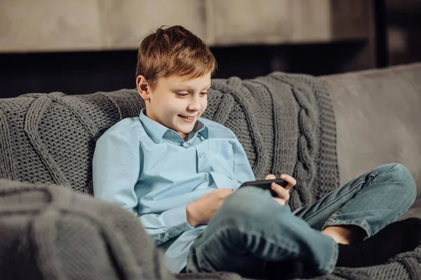Χαρούμενο αγόρι παίζει στο τηλέφωνο ενώ κάθεται στον καναπέ — Φωτογραφία Αρχείου