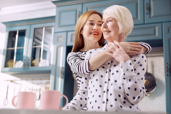 Mujer alegre abrazando a su madre mayor cerca del mostrador de la cocina —  Fotos de Stock