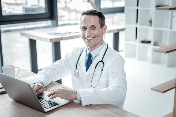 Satisfeito médico confiante sentado e usando o laptop . — Fotografia de Stock