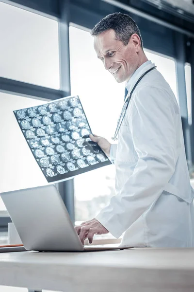 满意的愉快的医生看着笔记本电脑和持有 x 射线扫描. — 图库照片